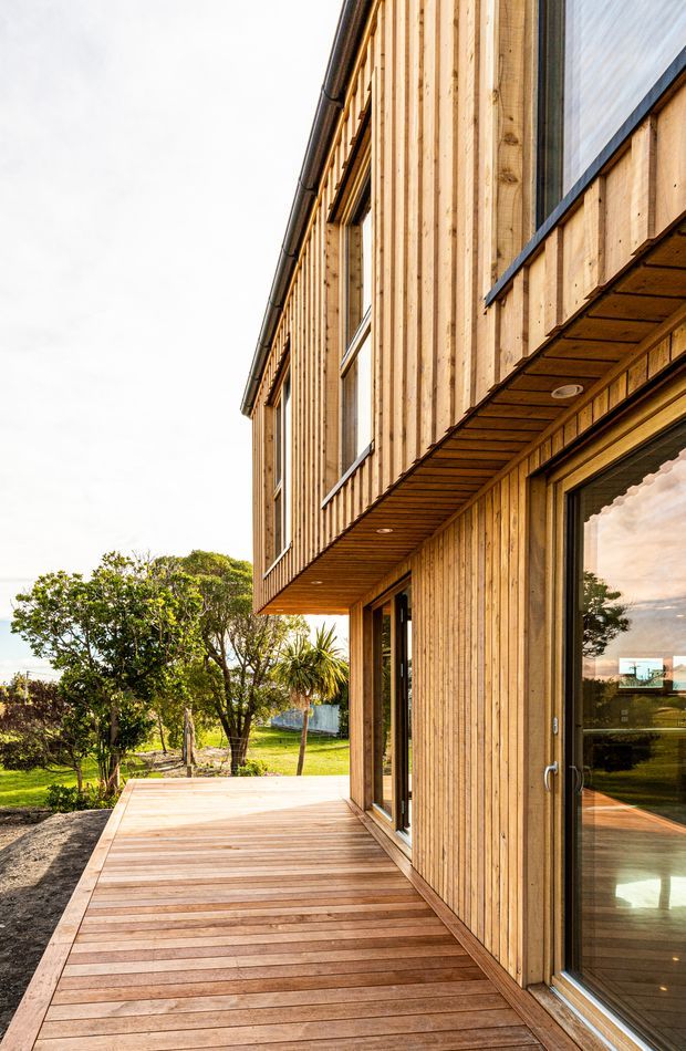 Exterior view of a contemporary two-storey building clad in vertical natural timber boards, with a projecting upper level and timber-lined soffit. Large dark-framed glazed doors open onto a broad timber deck, overlooking a landscaped garden. The image emphasizes the warm timber facade, deck, glazing, and cantilevered form.