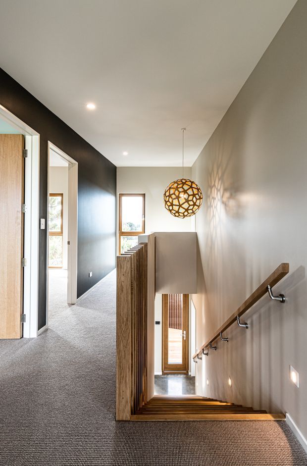 A bright, modern residential stairway viewed from an upper landing. The staircase descends alongside a pale wall with a timber handrail and black metal brackets, while a vertical timber balustrade screens the stair opening. Grey carpet covers the landing and stairs, and timber-framed windows and doors bring in natural light. A spherical woven pendant light casts a geometric shadow on the wall. A hallway and doorway are visible at the upper level.