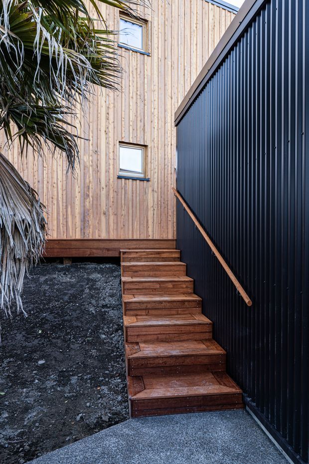 Exterior view of a compact stairway beside a contemporary building. The stairs and handrail are timber, leading between a vertically clad natural timber wall and a dark corrugated metal wall. A rough dark landscaped ground surface and tropical planting frame the access route, with small square windows visible in the timber-clad facade.