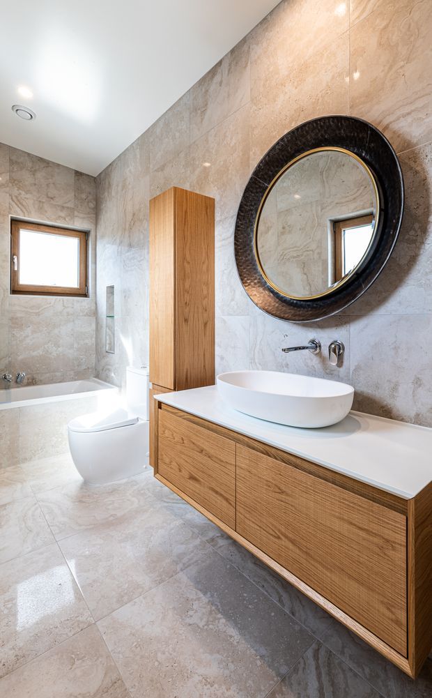 Bright contemporary bathroom with large beige stone-look wall and floor tiles, a floating natural timber vanity with white countertop and vessel basin, wall-mounted tapware, round framed mirror, freestanding toilet, bathtub, and timber-framed window. The room has clean lines, integrated storage, and a warm neutral palette.