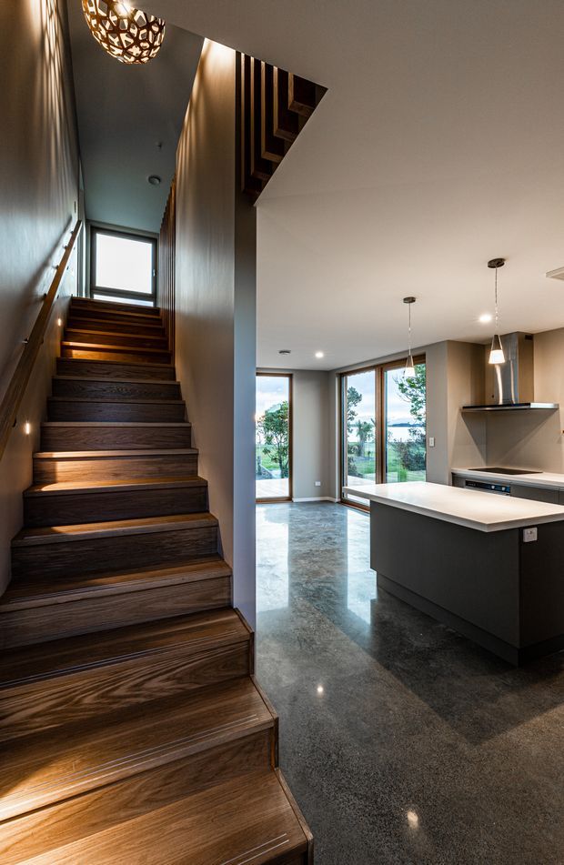 A contemporary open-plan kitchen and stairway interior. A dark timber staircase with integrated step lighting rises along the left side beneath a sculptural pendant light and open stair void. To the right is a minimalist kitchen with a large dark island and white benchtop, streamlined cabinetry, pendant lights, and a stainless-steel rangehood. Polished grey concrete flooring and expansive windows create a bright connection to the outdoors.