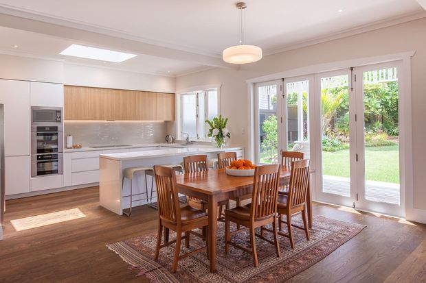 Bright, spacious open-plan kitchen and dining area with white cabinetry, a timber kitchen island, integrated appliances, and a long wooden dining table with matching chairs. Large white-framed glazed French doors open to a leafy garden, while a skylight and pendant light provide abundant natural and artificial illumination. The room combines warm timber flooring and furniture with light neutral finishes and traditional ceiling detailing.