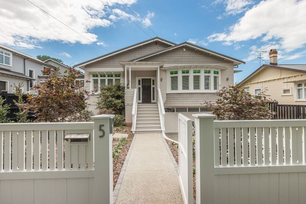 Front exterior view of a single-storey detached house with a prominent gabled roof, weatherboard cladding, enclosed front porch, raised entry stairs, and a central pedestrian path through a low pale fence and gate. The facade features a bay-like arrangement of multi-pane windows and restrained traditional detailing, with neighbouring homes visible on both sides.