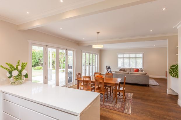Bright open-plan interior viewed from a white kitchen island, with a dining area and living room beyond. The space features warm beige walls, white cabinetry and trim, timber flooring, French doors opening to greenery, recessed lighting, and traditional decorative cornices. A wooden dining table and chairs sit on a patterned rug, while a neutral sofa occupies the living area.