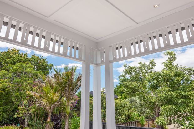 Bright covered outdoor veranda or enclosed porch with white painted posts, decorative vertical balustrade-style screening, and a recessed coffered ceiling. The open sides overlook lush tropical-subtropical garden planting and blue sky. The image focuses on the architectural detailing of the porch rather than furnishings.