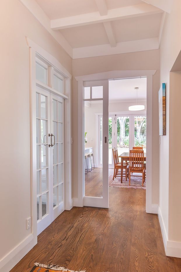 Bright residential hallway with warm timber flooring, pale painted walls, white trim, glazed French doors on the left, and a glazed doorway leading into a dining area. The corridor has a high exposed-beam ceiling, while the adjoining dining room contains a timber table and chairs, pendant lighting, and views to greenery outside.