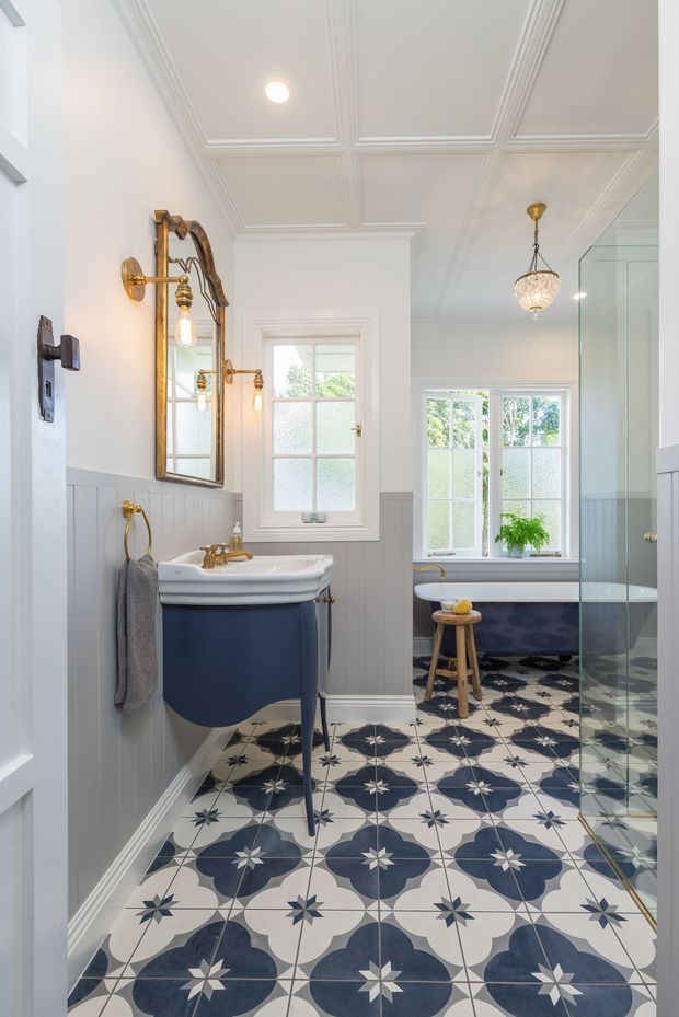 Bright, traditionally styled bathroom with a freestanding navy vanity, white basin and brass tapware, patterned blue-and-white floor tiles, white painted wall panelling, a decorative coffered ceiling, vintage-style mirror, bathtub, and glass shower enclosure. Frosted windows provide abundant natural light, while a crystal pendant adds a refined accent.