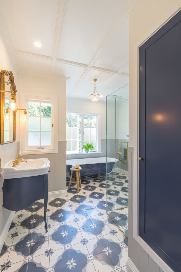 Bright, elegant traditional bathroom featuring a freestanding claw-foot bathtub, pedestal basin with dark navy painted base, glass-enclosed shower, decorative patterned floor tiles in blue, grey and white, white wall panelling, ornate mirror, polished brass fixtures, and a coffered ceiling. A dark navy paneled door occupies the foreground.