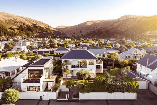 Aerial view of a leafy suburban residential neighbourhood set in a valley, with detached homes, landscaped gardens, white fences, and a mix of pitched-roof and contemporary houses. Steep, dry hills rise directly behind the settlement.
