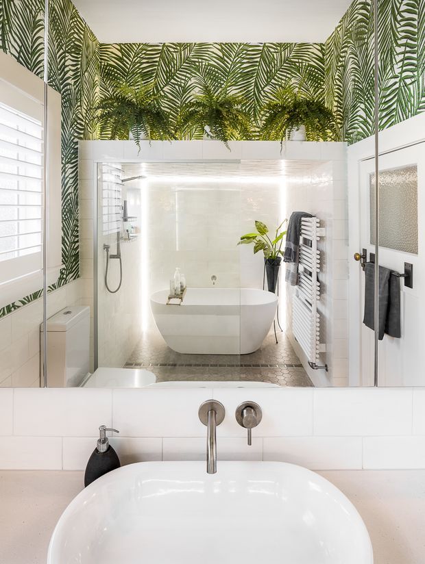 Bright contemporary bathroom featuring a freestanding white bathtub, walk-in shower, wall-mounted towel radiator, white tiled walls, pale stone or tile flooring, and a large mirrored vanity wall. Bold green tropical leaf wallpaper wraps the upper walls and is complemented by hanging plants. White plantation shutters and black and chrome fixtures add contrast.
