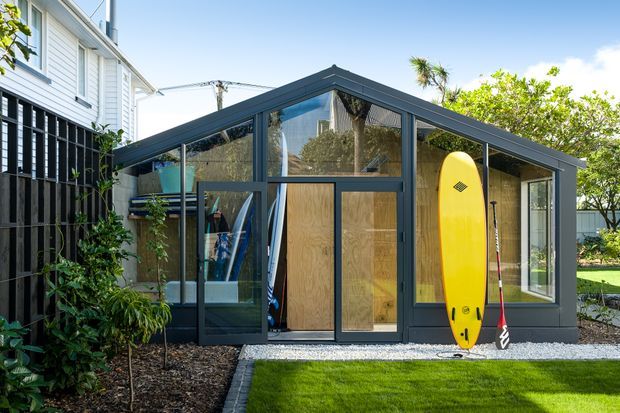 A compact contemporary backyard outbuilding or surf shed with a steep gabled roof, dark charcoal framing and cladding, and extensive glazing. Surfboards and paddles are stored inside and displayed outside, with a small landscaped lawn and gravel perimeter. A white weatherboard house is visible alongside it.