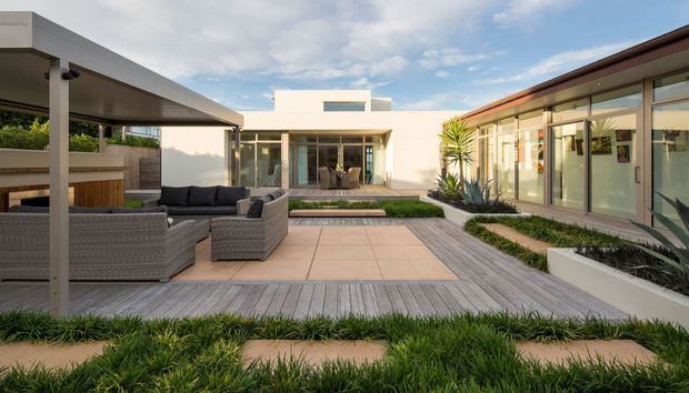 A contemporary single-storey courtyard residence arranged around a landscaped outdoor entertaining area. The image shows a paved patio with wicker outdoor seating, a covered outdoor structure, timber-look decking, raised garden beds, extensive glazing and low-profile flat-roofed buildings.