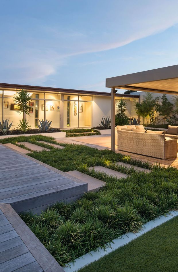 A landscaped modern courtyard viewed at dusk, featuring a single-storey flat-roofed residence with extensive floor-to-ceiling glazing, warm interior lighting, sculptural agave and yucca plantings, grass-filled garden beds, concrete stepping paths, and a timber deck. A covered outdoor lounge with woven seating occupies the right side of the courtyard.