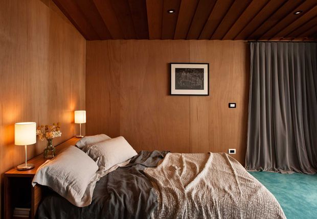 A warmly lit bedroom with full-height timber wall panelling and a matching exposed timber slat ceiling. The room contains a large bed with layered neutral bedding, bedside tables and lamps, framed artwork, and full-height charcoal-grey curtains. A small area of teal carpet is visible at the foot of the bed. The mood is intimate, warm, and refined.