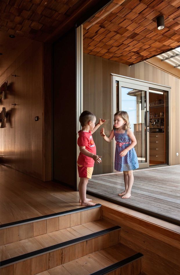 Interior view of a warm timber-lined transition space with broad stepped stair seating leading to a raised timber deck. Two children stand near a large sliding glass door, with a built-in timber-clad room and shelving visible beyond. The ceiling features exposed small timber battens or shingles, while daylight enters through the glazed opening and a translucent roof section.