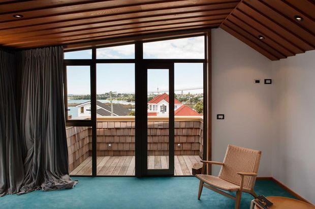 A compact, refined interior room with a dramatic slatted timber ceiling, teal carpet, white walls, dark-framed floor-to-ceiling glazing, and a glazed door opening to a small timber-clad balcony. A woven timber lounge chair sits in the foreground beside full-height charcoal curtains. The view beyond shows neighbouring homes and a distant urban/coastal setting.
