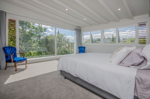 Bright, spacious bedroom with a large upholstered bed, white bedding, grey carpet, and extensive windows. The room features white plantation-style louvred windows, a broad glazed wall, and a white coffered or exposed-beam ceiling. Two vivid blue upholstered occasional chairs provide colour accents, while leafy greenery is visible outside.