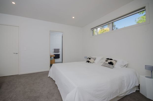 Bright, minimalist bedroom with a large white bed, white bedding and pillows, grey carpet, white walls, recessed ceiling lights, narrow high-set windows, and a simple white bedside table with lamp. A partially visible vanity or storage area appears through the open doorway.
