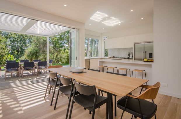 Bright open-plan dining and kitchen interior with a long timber dining table, mixed dining chairs, white cabinetry, and a stone-look splashback. Large glazed folding or sliding doors open to a covered outdoor dining terrace, while linear roof louvres cast strong striped shadows across the timber floor. The space has a clean contemporary fit-out with abundant natural light and a leafy outlook.