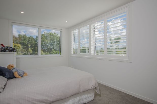 Bright, compact bedroom with a neatly made bed, pale bedding, white walls and trim, recessed ceiling lights, carpeted floor, and extensive corner glazing. The windows include white plantation shutters, with leafy greenery visible outside. A few toys sit on the left windowsill.