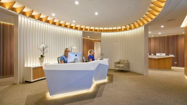 A refined contemporary commercial reception area with a sculptural white reception desk, two staff workstations, and a curved feature wall of vertical white slats. Warm concealed lighting and repeated gold-toned geometric ceiling fins create a softly illuminated circular canopy. The space includes warm timber reception joinery, vertical timber accents, neutral carpet flooring, recessed ceiling lights, and a secondary counter area in the background.