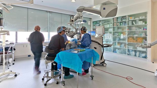 Bright, modern clinical operating or procedure room with medical staff gathered around a draped procedure table. The space features white seamless flooring, pale walls, extensive glazed cabinetry displaying medical supplies, large medical equipment, ceiling-mounted surgical lights, and generous daylight through frosted glazing.