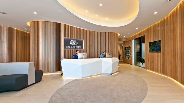 Contemporary medical eye institute reception area with a sculptural white angular reception desk, two workstations, curved timber slat wall panelling, warm integrated cove and strip lighting, pale timber-look flooring, and a large rounded ceiling feature. A lounge chair and indoor planting are visible at the edges of the space.