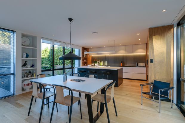 Open-plan contemporary kitchen and dining area with pale timber flooring, white walls and cabinetry, a dark island bench, stainless-steel splashback, extensive black-framed glazing, and a large white dining table surrounded by modern timber and metal chairs. Built-in shelving and a sculptural pendant light add visual interest, while a blue lounge chair sits near the glazing.