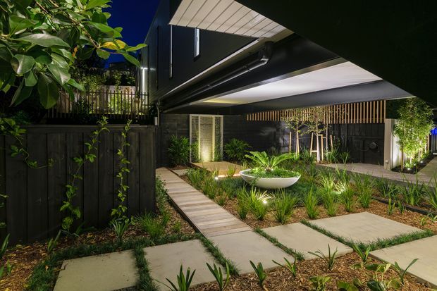 A landscaped courtyard and entry garden at night, viewed beneath a dramatic cantilevered upper structure. Dark vertical cladding and fencing contrast with illuminated pale soffits, timber-like screening, concrete stepping slabs, a narrow timber walkway, and layered ornamental planting. A sculptural white bowl planter forms a focal point in the garden.