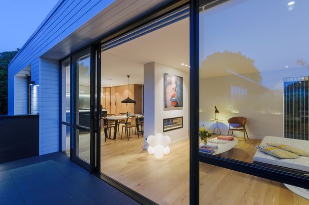Evening view through large black-framed glazed sliding doors into a contemporary open-plan interior. The room features pale timber flooring, white walls, warm timber cabinetry, a dining table with pendant light, a small living area, and an adjacent bedroom visible through the glazing. The exterior has horizontal light-coloured cladding and a dark paved terrace.
