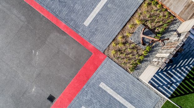 Aerial view of a contemporary landscaped outdoor area featuring geometric paving zones, dark grey herringbone pavers with contrasting light strips, broad charcoal surfacing edged in vivid red, planted garden beds with gravel, timber stepping planks, and a partially visible striped shade structure.