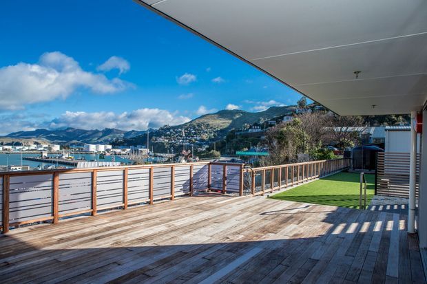 A spacious elevated timber deck or terrace with horizontal privacy screening and timber-framed glass balustrades, extending beneath a broad roof overhang. The outdoor space overlooks a marina, waterfront settlement, and mountainous hills. Bright daylight, weathered timber decking, and open views create a contemporary coastal setting.