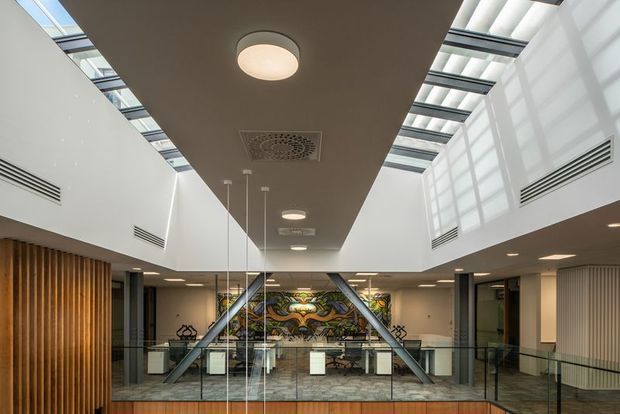 Interior view of a spacious contemporary multi-level commercial atrium or lobby. The space features a large central ceiling bulkhead with recessed circular lights, extensive clerestory glazing and skylights, white walls, exposed steel structural braces, glass balustrades, timber vertical wall screening, and a decorative mural or artwork at the far end. The open void and upper-level circulation create a bright, airy institutional or commercial environment.
