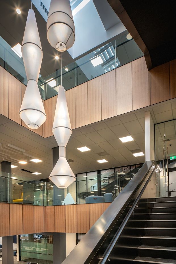 A multi-level contemporary commercial interior atrium with a prominent staircase, glass balustrades, timber-lined walls, suspended sculptural pendant lights, and a visible upper-level walkway or mezzanine. The space has a bright, polished, institutional or office-like fit-out with extensive glazing and a high void.