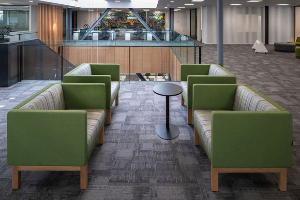 A contemporary commercial interior showing a lounge and circulation area. Four green upholstered armchairs and striped sofas are arranged around a small round black table on grey patterned carpet. A glazed balustrade overlooks a lower level, with timber and glass partitions, a colourful decorative artwork, and a large open office or reception area in the background.