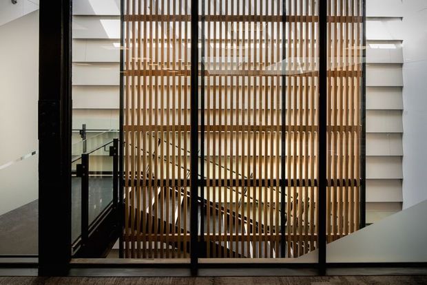 Interior architectural view of a multi-level stairway or atrium enclosed by black-framed glazing and a prominent vertical timber slat screen. White walls, horizontal architectural fins, glass balustrades, and a dark polished floor create a modern, carefully detailed circulation space.