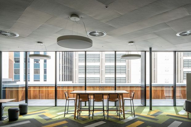 Contemporary commercial interior with a shared dining or collaboration area positioned beside full-height windows. The room features a long timber table with bar-height chairs, built-in timber wall panelling, patterned geometric carpet, large circular pendant lights, exposed acoustic ceiling tiles, and views of neighbouring urban buildings.