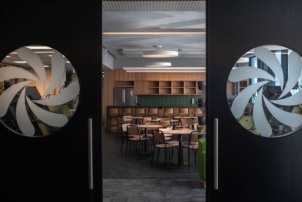 View through open black double doors into a contemporary commercial café or dining space. The doors feature large circular decorative glass panels with swirling frosted patterns. Inside are rows of small tables and black-framed chairs, a long green-tiled counter or splashback, warm timber shelving and cabinetry, dark carpet flooring, and a prominent industrial-style exposed ceiling with linear and circular pendant lighting.