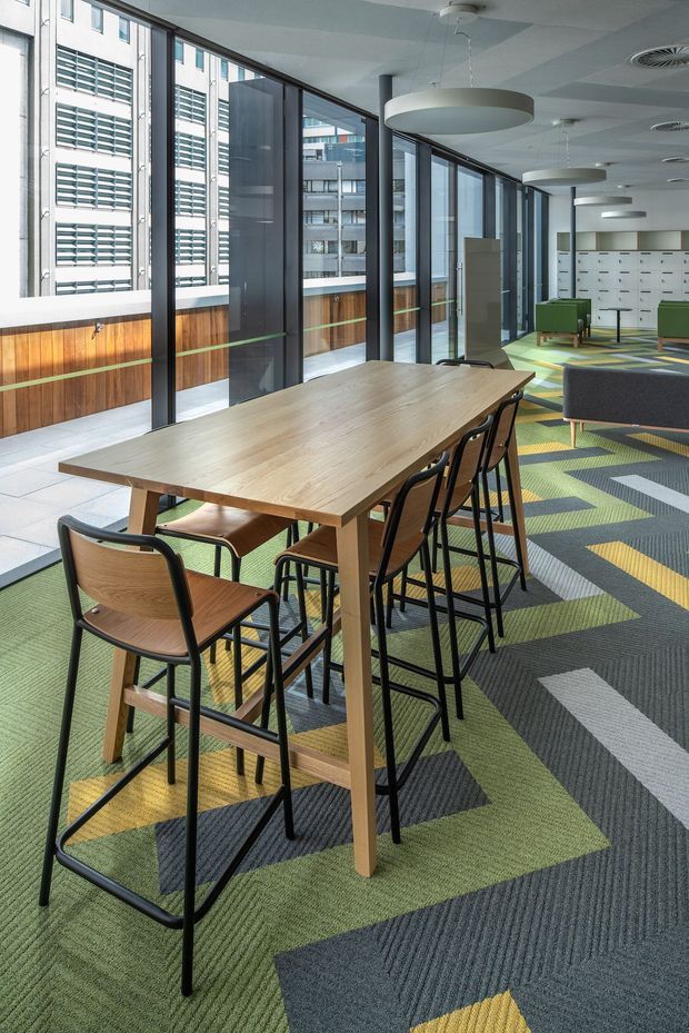 A bright contemporary commercial interior, likely a workplace breakout or collaboration area, with a long timber communal table and black-framed timber stools in the foreground. Full-height glazed windows line the left side, overlooking adjacent urban buildings. The space includes patterned modular carpet in green, charcoal, yellow and pale blue, green upholstered lounge chairs, large circular pendant lights, exposed ceiling services, and built-in white storage or locker units at the far end. Timber wall panelling with a green horizontal accent adds warmth to the otherwise modern office fit-out.