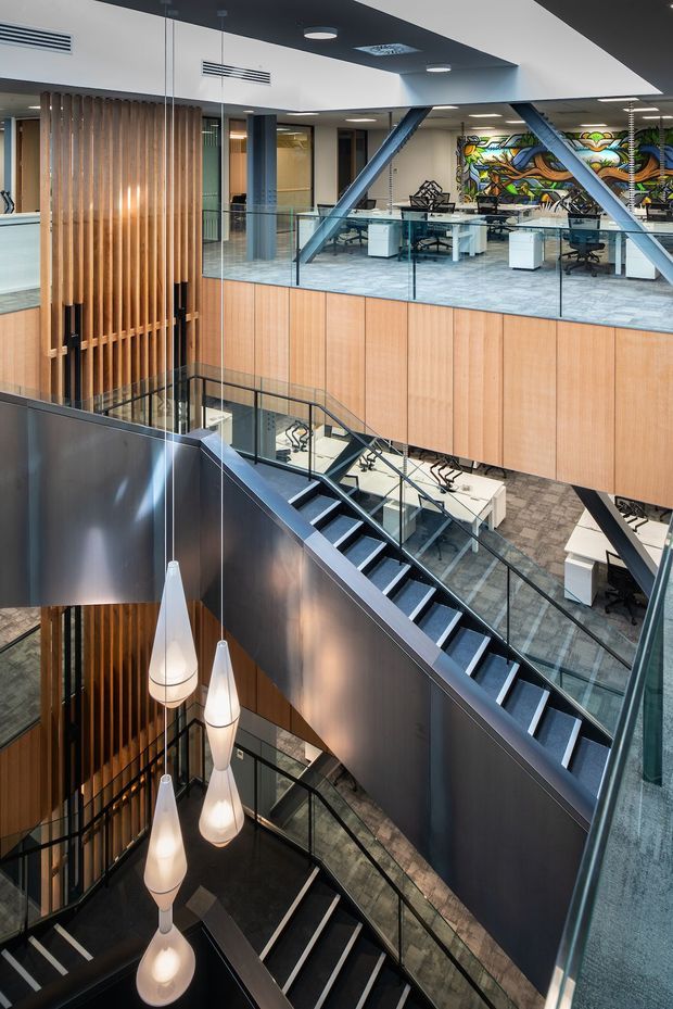 A contemporary multi-level commercial office atrium viewed from an upper landing, featuring dramatic intersecting staircases with dark metal stringers, glass balustrades, carpeted steps, timber slat screens, warm timber wall panelling, dark feature walls, exposed structural bracing, and a series of pendant lights descending through the void. Open-plan workspaces with desks and chairs are visible across the atrium, along with a colourful mural on the far wall.