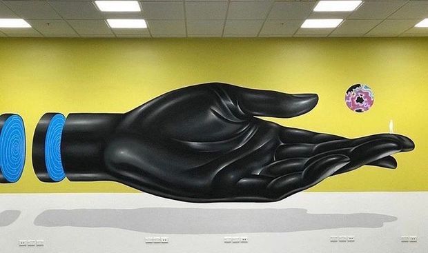 Interior wall display featuring a large glossy black sculptural artwork resembling an abstract flowing hand or wing, mounted against a vivid yellow wall. Blue circular forms appear at the left edge, with a small colourful circular artwork above the right side. The space has a suspended acoustic tile ceiling, fluorescent-style rectangular lights, and a pale lower wall with electrical outlets.