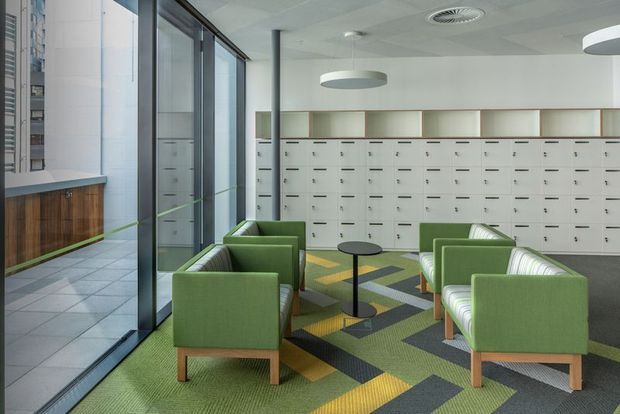 Bright contemporary commercial interior, likely a reception or shared office lounge, with green upholstered armchairs arranged around small round tables. A large wall of white built-in locker or filing units spans the rear, while full-height glazing overlooks an adjacent exterior walkway. The space has a patterned geometric carpet in green, grey, yellow, and black, pale acoustic ceiling tiles, suspended circular lights, and a timber-fronted built-in counter along the window wall.