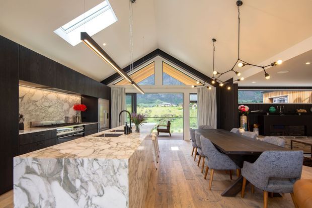 Open-plan kitchen and dining area with dark timber cabinetry, a large veined marble island, warm timber flooring, and a pitched glazed gable opening to a green landscape. Contemporary pendant and track lighting illuminate the space, with a dining table and upholstered chairs positioned beside the kitchen.