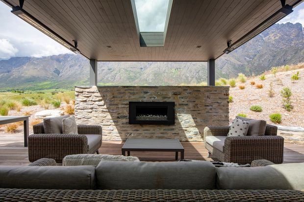 A covered outdoor lounge overlooking a dramatic mountainous landscape. The space features a low stone-clad fireplace wall with a built-in black fireplace, timber decking, a timber-lined ceiling with a central skylight, dark structural posts, and woven outdoor seating arranged around a coffee table.