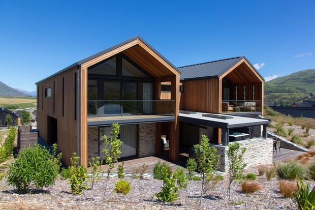 Exterior view of a contemporary alpine-style residential property composed of two connected gabled volumes. The building features dark vertical timber cladding, large black-framed glazing, exposed timber-lined gable soffits, balconies and terraces, stone masonry walls, and dark metal pitched roofs. It sits in a landscaped sloping site with mountain scenery in the background.