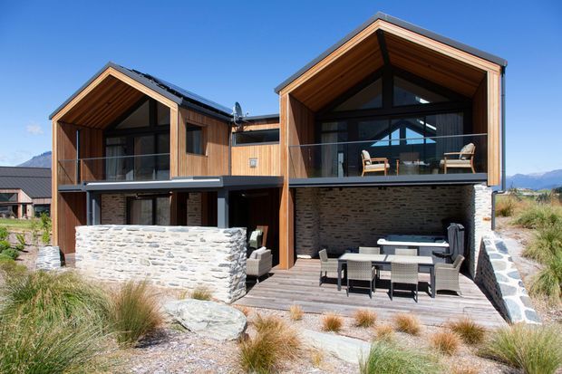 Exterior view of a contemporary two-storey alpine residence with two prominent timber-clad gabled volumes, extensive dark-framed glazing, recessed balconies, and exposed timber soffits. Rough stone masonry forms the lower-level walls and terrace edging, while a timber deck with outdoor dining and lounge furniture opens onto a landscaped, dry-grass setting with mountains in the distance.