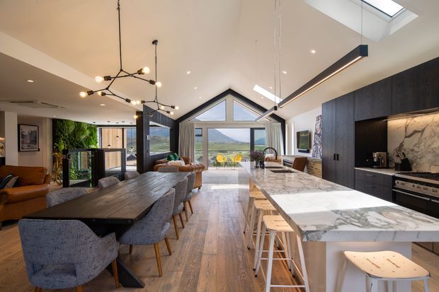 Open-plan contemporary kitchen, dining and living area with a high vaulted ceiling and dramatic glazed gable end overlooking a scenic landscape. The space features a large marble-look island with bar stools, dark timber cabinetry, timber flooring, a substantial dining table, upholstered dining chairs, and modern sculptural pendant lighting. Skylights and extensive glazing create a bright, refined interior.