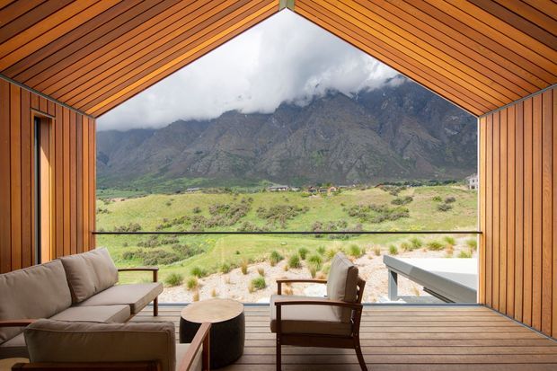 A timber-lined covered outdoor living space frames a dramatic mountain landscape through a large open elevation. The space features warm vertical and slatted timber surfaces, a timber deck, a neutral upholstered sofa, lounge chairs, and a small round coffee table, creating a sheltered viewing and entertaining area.