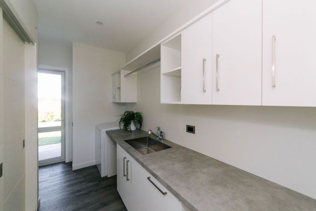 Compact, bright white galley-style utility kitchen or laundry space with extensive white built-in cabinetry, a long grey concrete-look benchtop, integrated double sink, dark timber-look flooring, and a glazed exterior door at the end. The simple clean-lined fit-out has minimal decoration and abundant natural light.