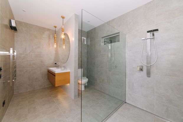 Contemporary bathroom with full-height light grey stone-look wall and floor tiles, a frameless glass shower enclosure, wall-mounted rain shower and handheld fixture, recessed shower niche, floating timber vanity with white basin, large round mirror, and warm pendant lighting.
